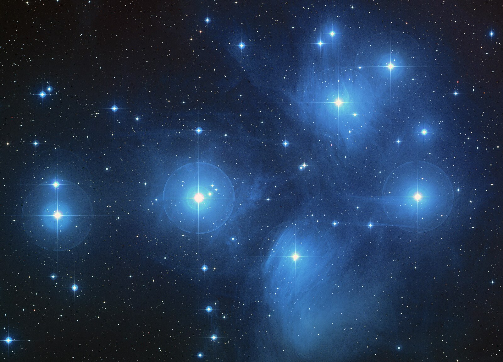 The Pleiades star cluster glowing blue with wispy reflection nebulae against a dark star field.